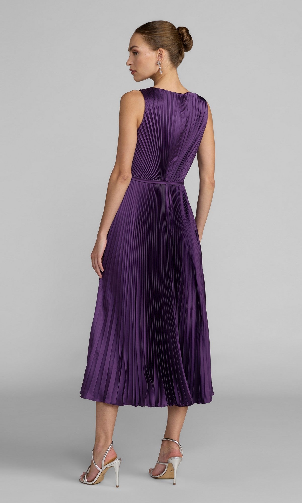 A woman stands with her back to the camera against a plain gray background, wearing the Midi Party Dress: Mac Duggal 56326—a sleeveless, pleated charmeuse dress featuring an A-line skirt in a rich purple hue—paired with silver heels.